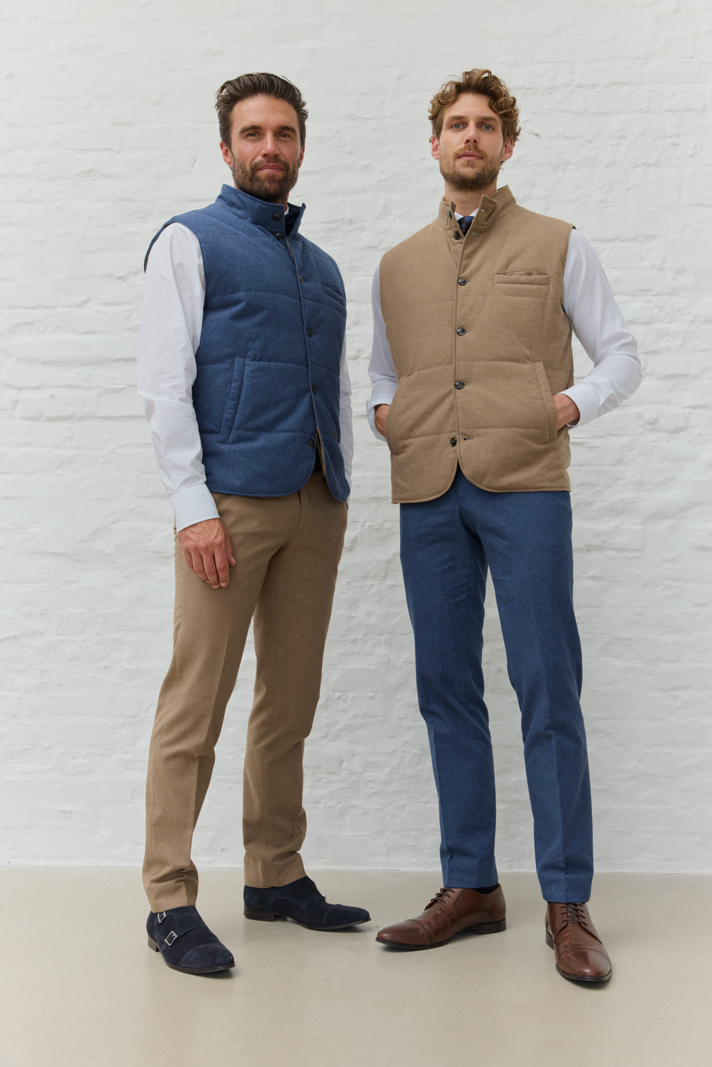Zwei Männer in Westen und Hosen vor weißer Wand.