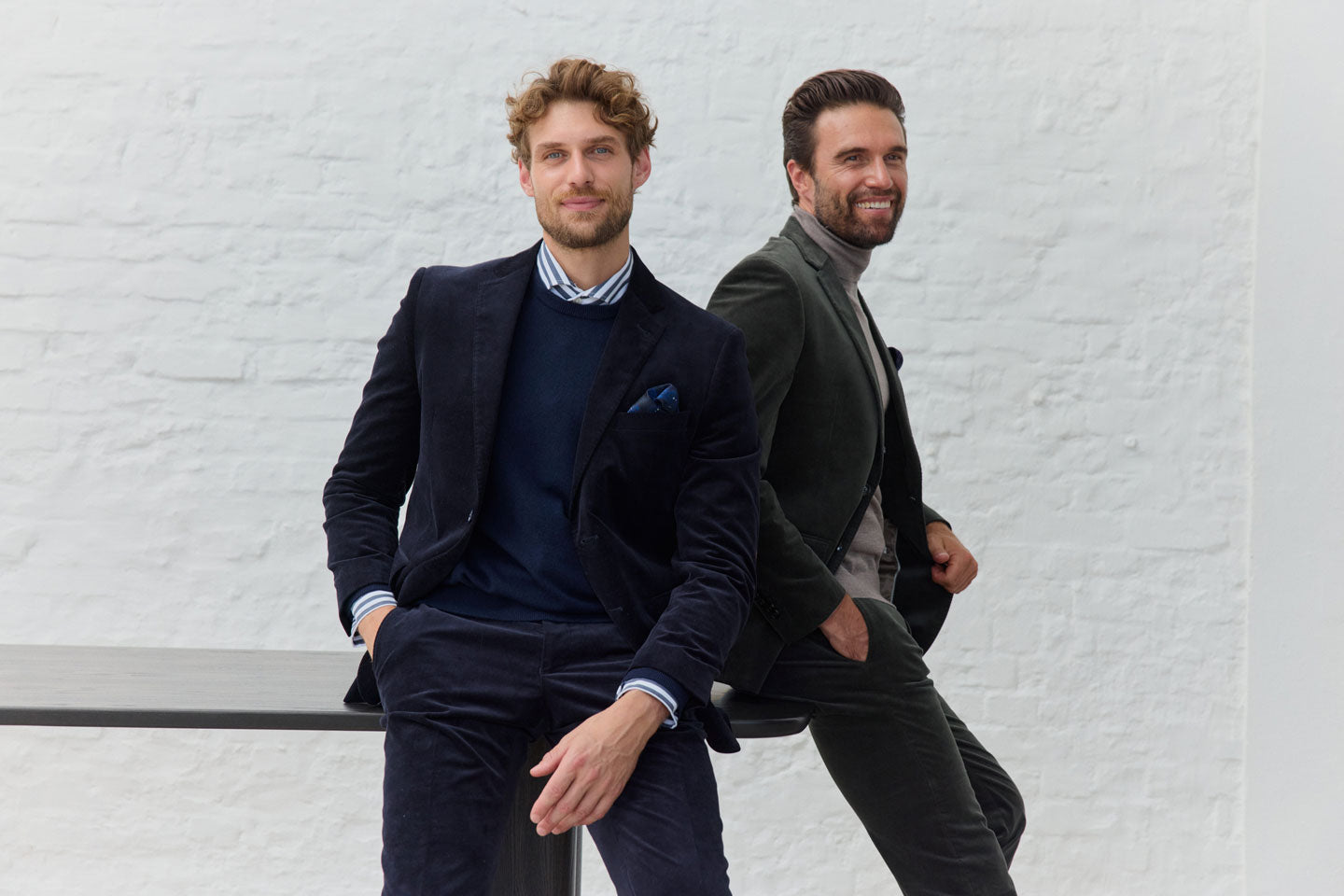 Zwei Männer in Anzügen vor weißer Wand.