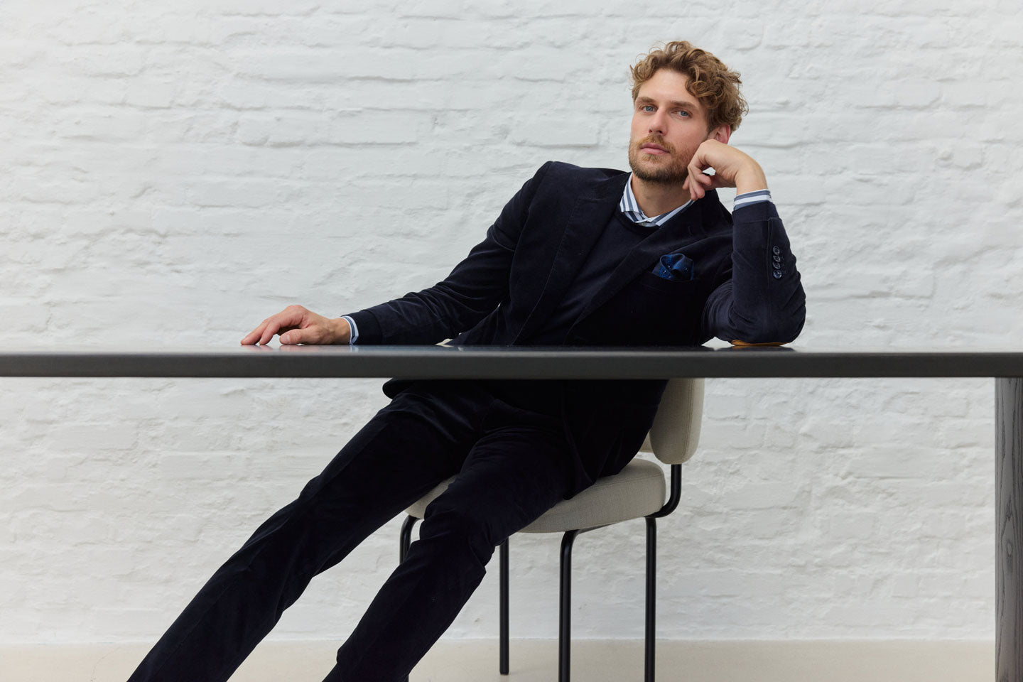 Mann in Anzug lehnt sich auf Tisch, weiße Ziegelwand.