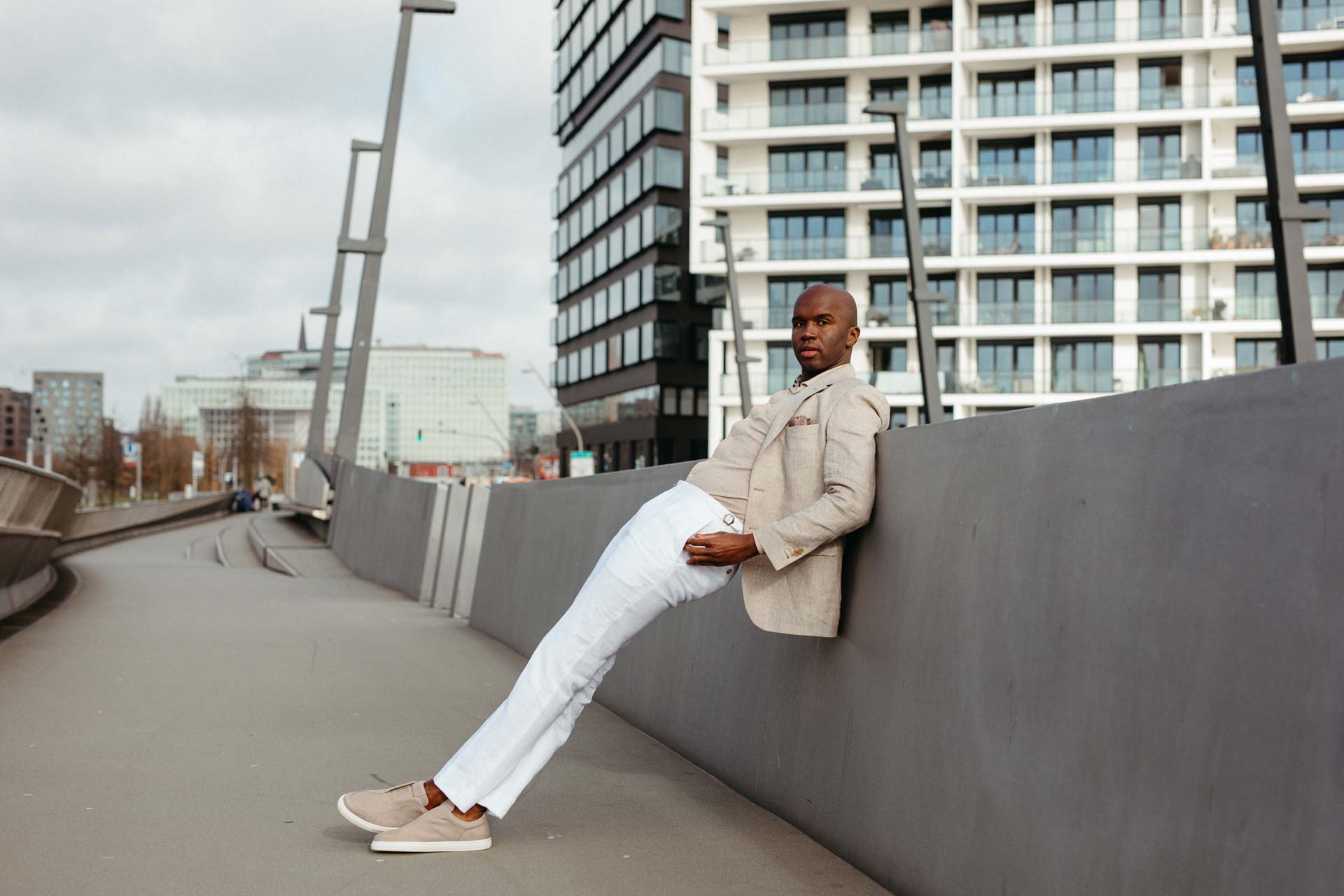 Mann in beiger Jacke und weißer Hose lehnt draußen.